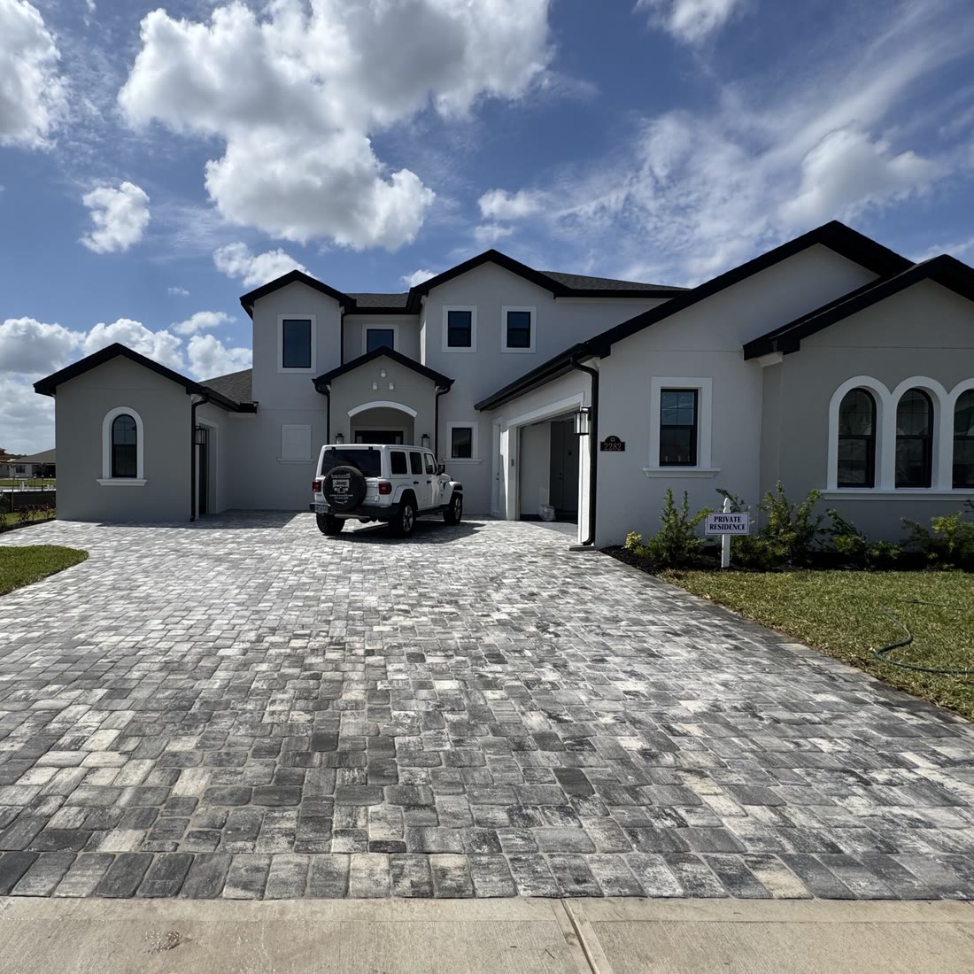 A grey paver driveway in front of a two-storey white house with arched windows, a Jeep parked on it.