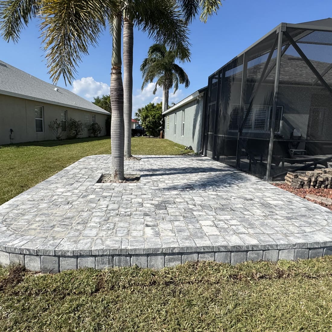 A raised paver pad built around two palm trees beside a screened pool.