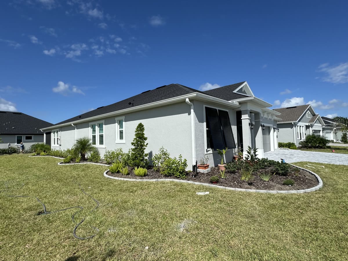 A long paver border along the front beds of a grey house, a wide lawn in front.