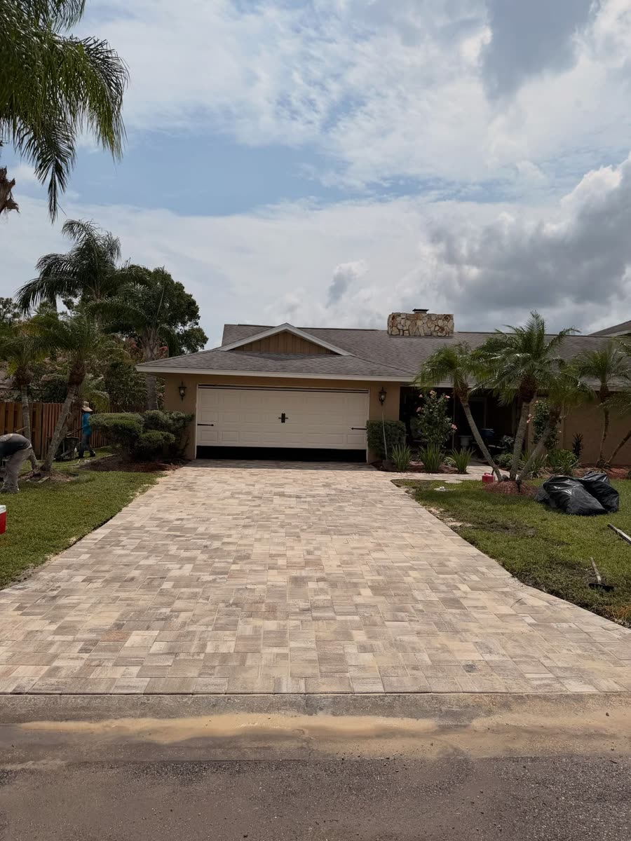 A long paver driveway in white, tan and charcoal leading to a garage, tall palms beside it.