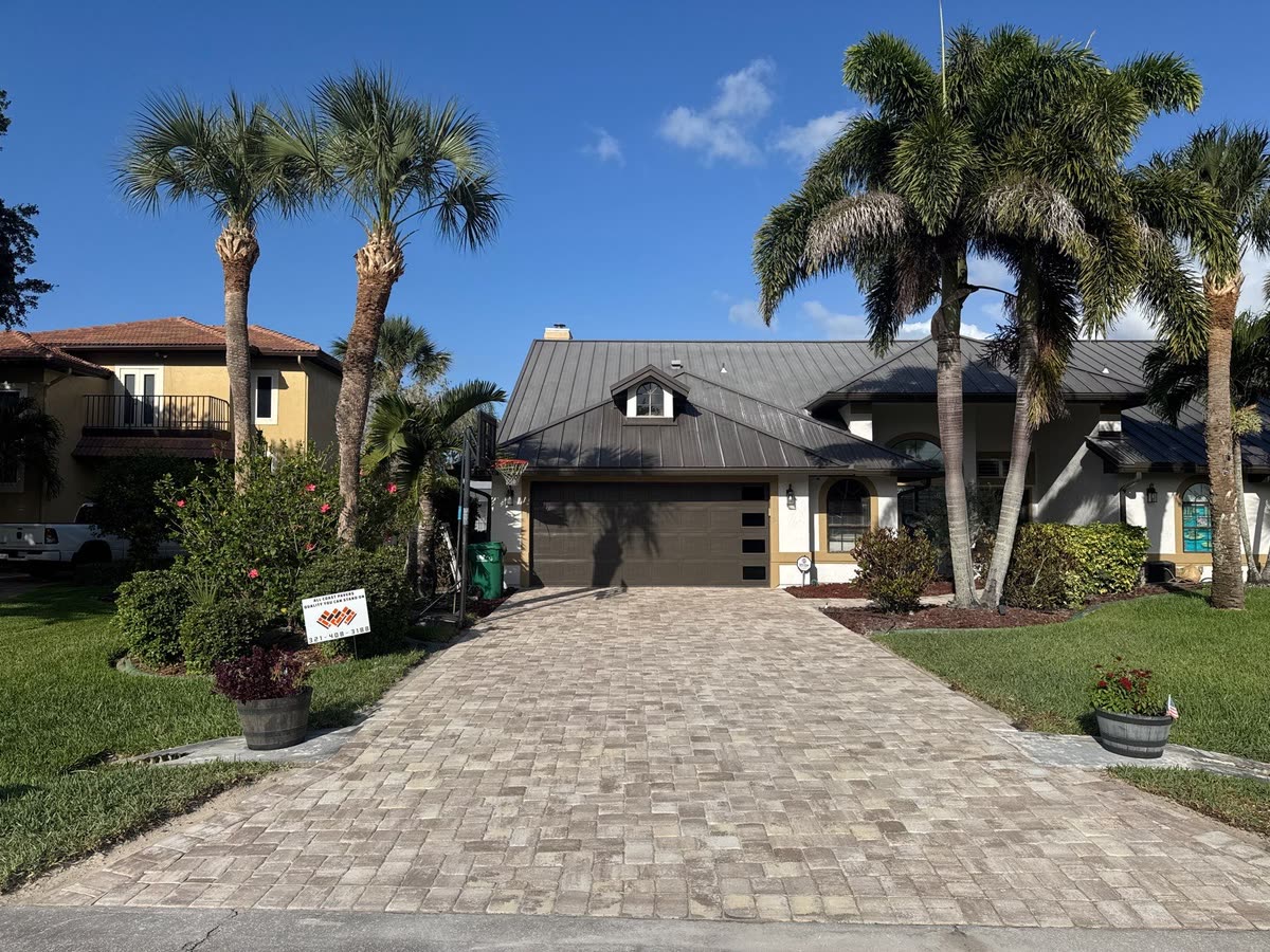 A long paver driveway lined with tall palms, leading to a grey house.