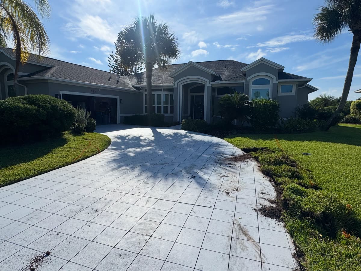 A very wide, pale paver driveway curving up to a house, palms along the lawn.