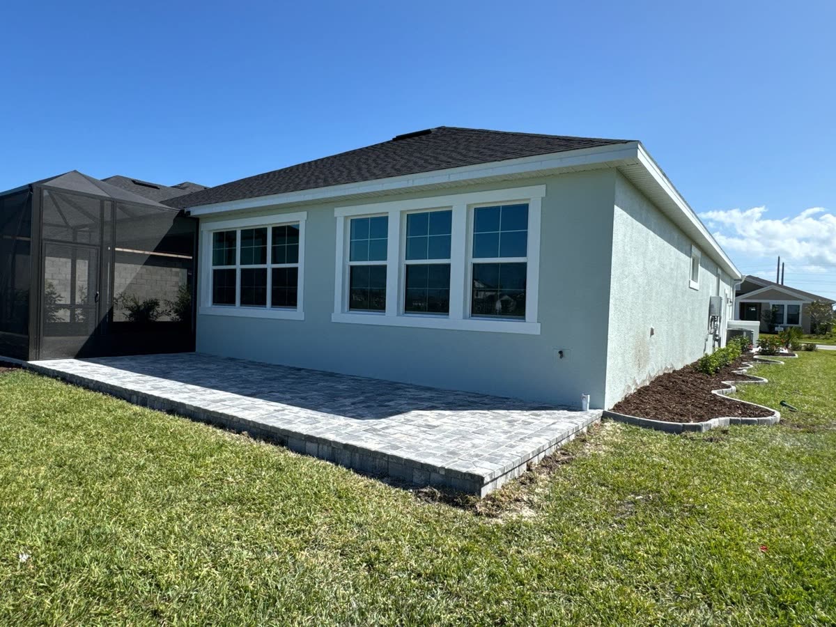 A paver patio across the back of a pale blue house, edged against the lawn.