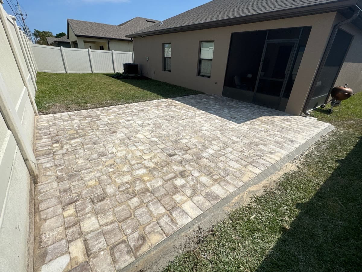 A paver patio behind a grey house, running up to a white fence.