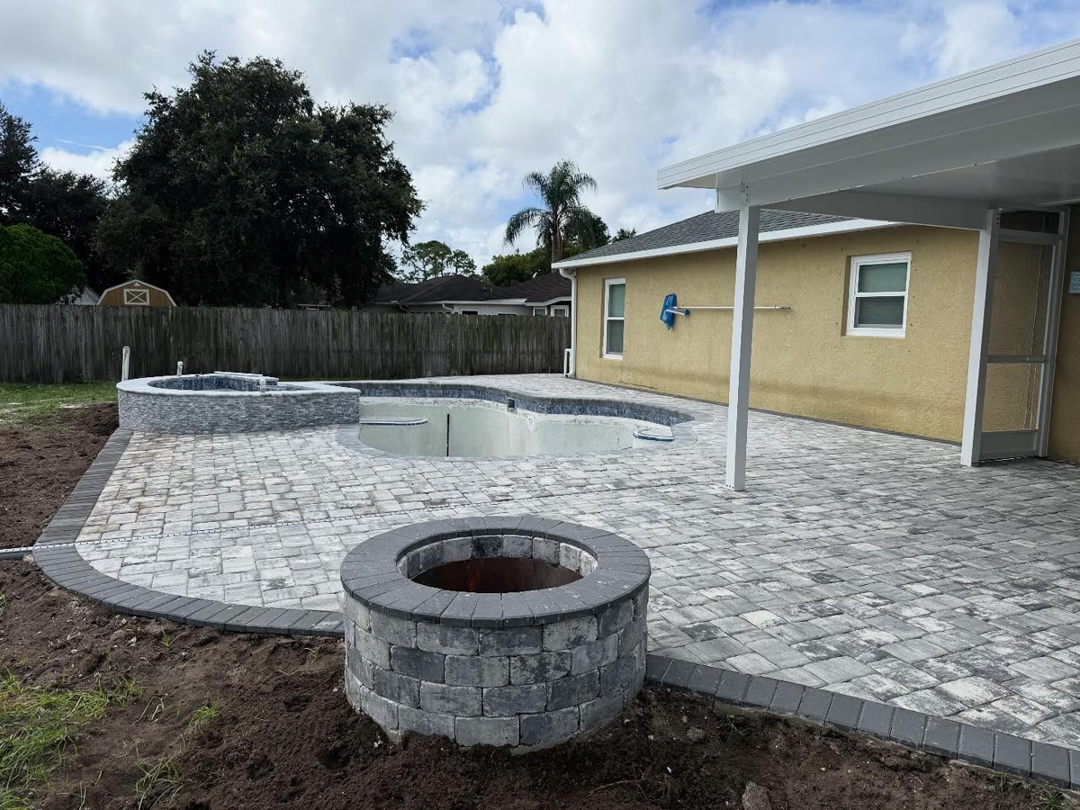 A paver pool deck with a round stone fire pit in front of a yellow house.