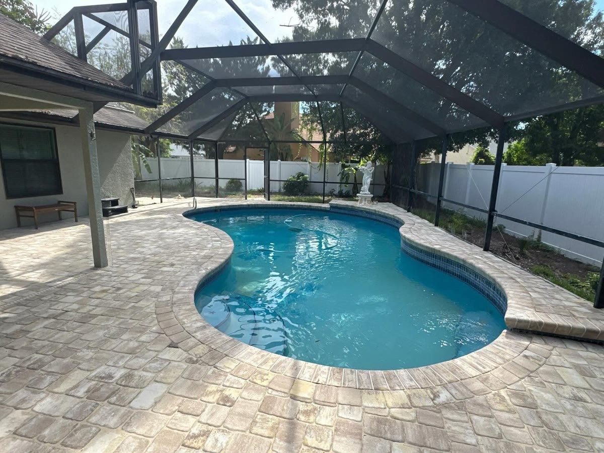 A kidney-shaped pool inside a screen enclosure, a grey paver deck all round it.