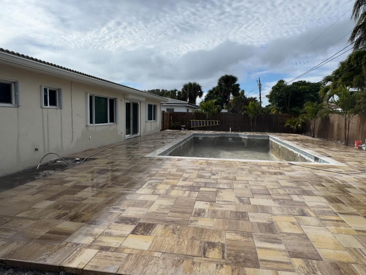 A new pool with a wide deck of tan and brown pavers, a wood fence around the yard.