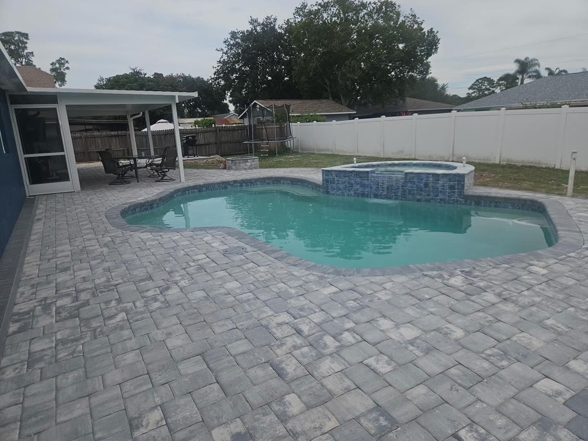 A pool and raised spa surrounded by a grey paver deck, a white fence behind.