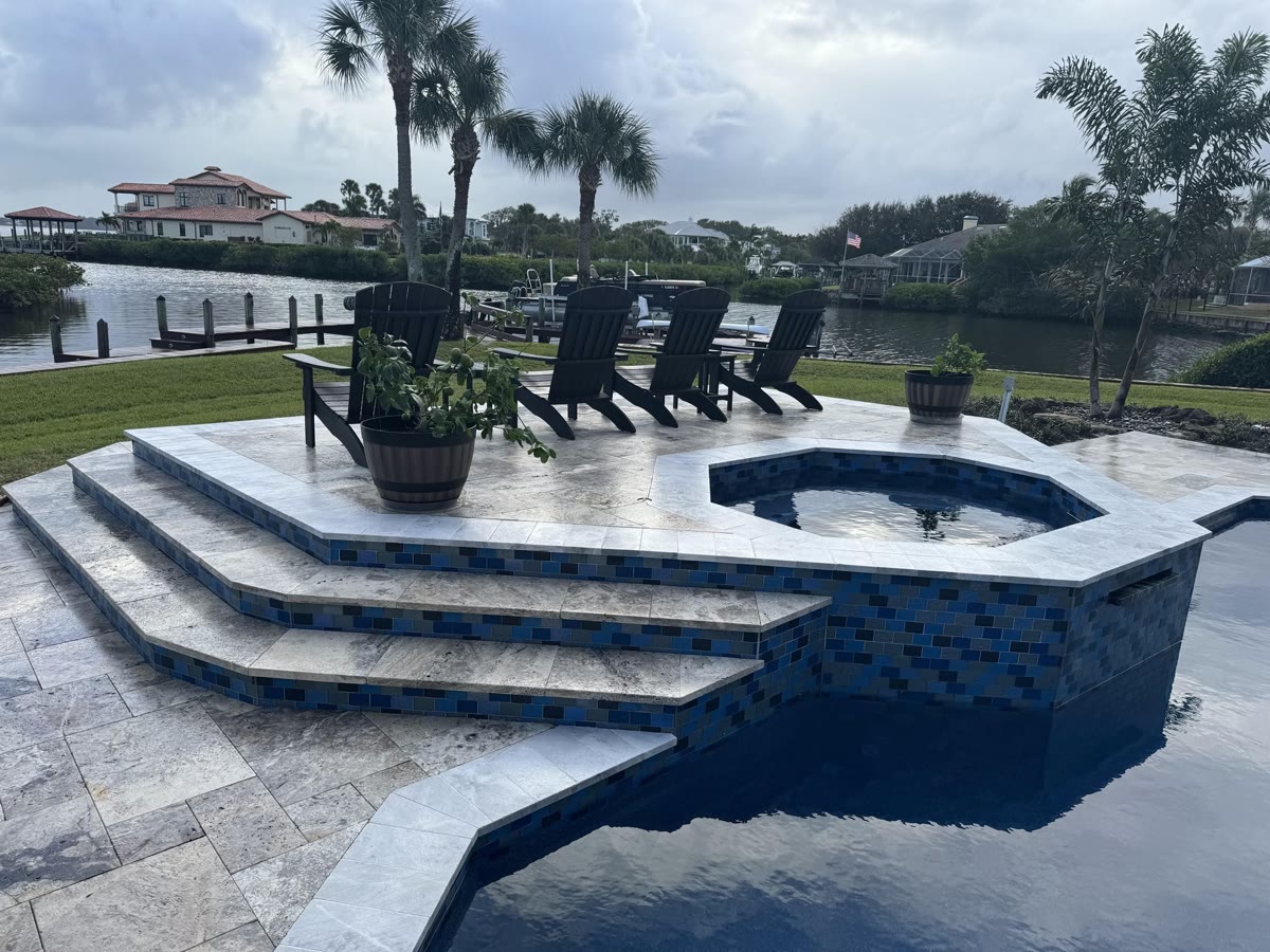 A waterfront pool and raised spa with a pale paver deck and steps, lounge chairs facing the water.